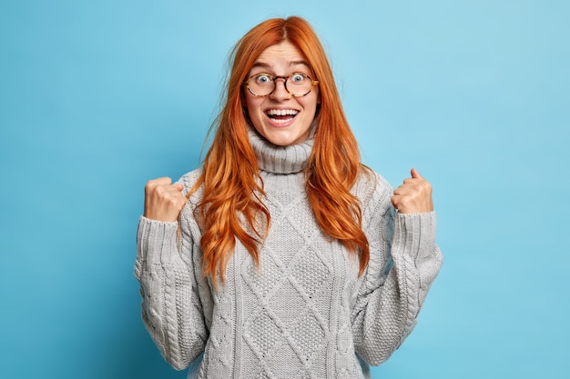 Бесплатное фото Радостная рыжая молодая женщина с веселым выражением лица радуется успеху, сжимает кулаки после достижения цели, одетая в вязаный серый свитер.