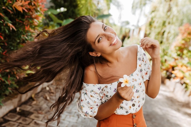 Free photo lady with white flower in her hand plays with hair woman in bright summer outfit with smile posing against background of plants