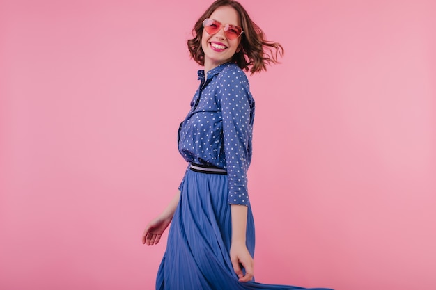Free photo laughing carefree girl in midi skirt enjoying indoor photoshoot. european female model in stylish blouse and sunglasses standing on pink wall.