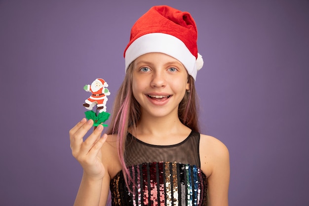 Free photo little girl in glitter party dress and santa hat showing christmas toy looking at camera with smile on face standing over purple background