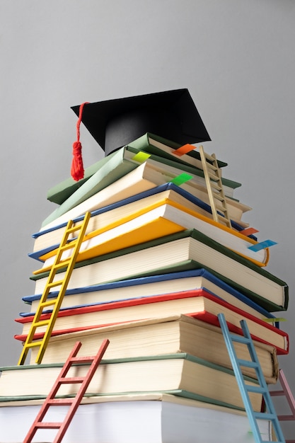 Бесплатное фото Низкий угол сложенных книг, выпускной колпак и лестницы для дня обучения