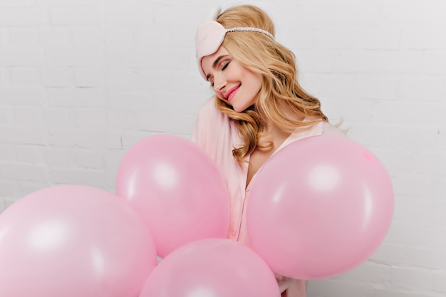Бесплатное фото Великолепная молодая женщина в розовой маске для глаз позирует с удовольствием, держа в руках гелиевые шары. крытый портрет потрясающей девушки, празднующей свой день рождения утром.