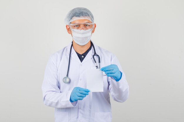 Male doctor in protective clothes holding white paper card and looking careful