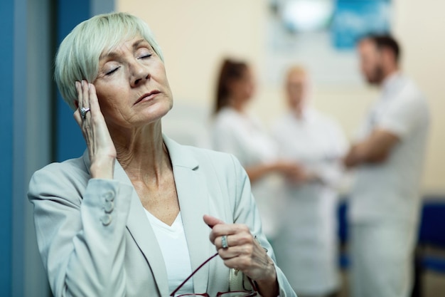 Зрелая женщина с закрытыми глазами держит голову от боли во время головной боли в коридоре больницы