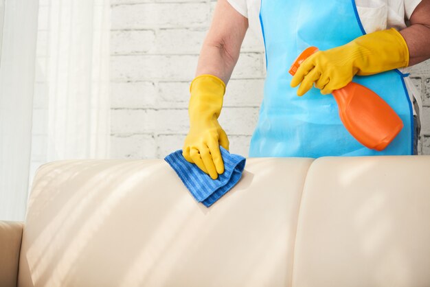 Средняя часть неузнаваемой домохозяйки вытирает кожаный диван спреем для полировки кожи