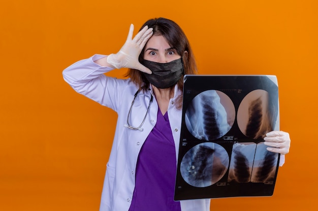 Бесплатное фото Женщина-врач средних лет в белом халате в черной защитной маске для лица и со стетоскопом, держащим рентгеновский снимок легких, выглядит удивленно с рукой возле головы, стоящей на оранжевом фоне