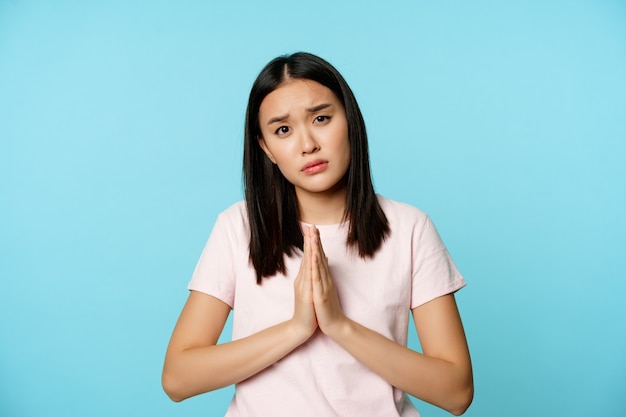 Бесплатное фото Несчастная азиатская девушка просит о помощи, умоляет, умоляет о чем-то, стоит грустно и мрачно на синем фоне.