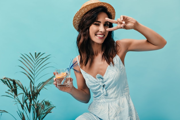 Бесплатное фото Симпатичная девушка показывает знак мира, подмигивает и держит воду с апельсином. улыбающаяся женщина с короткими темными волосами позирует возле небольшой пальмы.