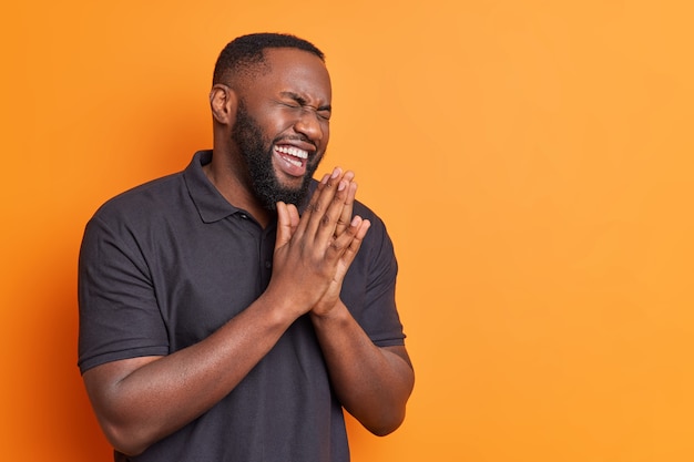 Free photo overjoyed bearded adult man rubs palms and laughs out happily dressed in casual black t shirt hears funny joke poses against bright orange wall with copy space for your text