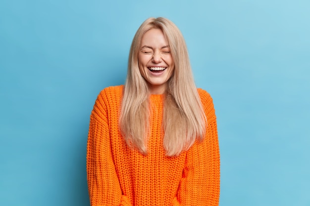 Бесплатное фото Обрадованная женщина с длинными светлыми волосами смеется, слышит что-то смешное, закрывает глаза, показывает белые зубы, носит оранжевый вязаный свитер