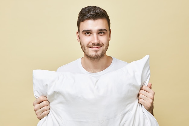 People, bedding and rest concept. Portrait of attractive happy young male with stubble posing isolated holding white pillow going to sleep, with positive smile, saying Good night