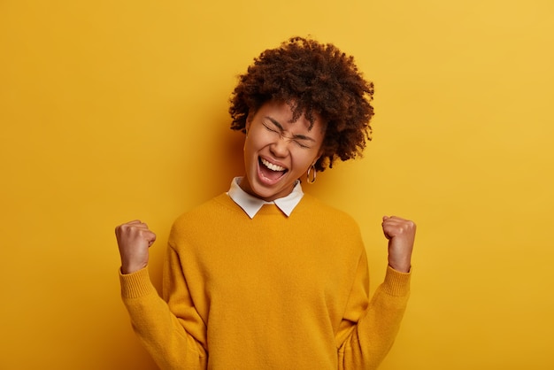 Free photo photo of cheerful curly haired woman feels like winner, clenches fists, makes victory gesture, exclaims with happiness, wears yellow jumper, achieves goal, gets triumph, poses indoor. yes, i did it!