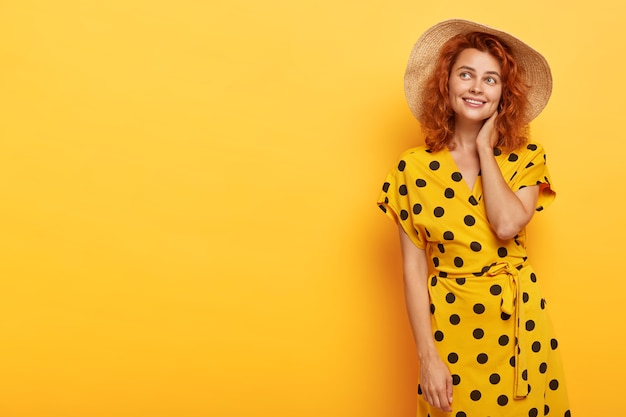 Бесплатное фото Фотография мечтательной красивой рыжей женщины, позирующей в желтом платье в горошек и соломенной шляпе