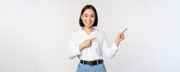 Foto gratuita ritratto di bella donna asiatica commessa che punta il dito a destra e mostra banner informativo o annuncio di vendita logo in piedi su sfondo bianco