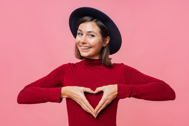 Free photo portrait of beautiful brunette woman in hat , shows heart gesture over chest, being passionate, express love to close person, stands.