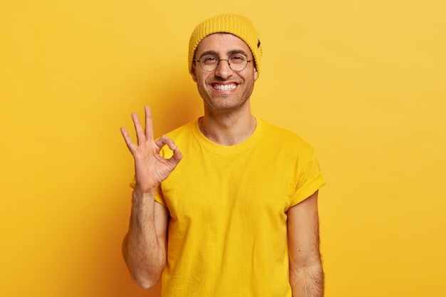 Free photo portrait of handsome young man makes okay gesture, demonstrates agreement, likes idea, smiles happily, wears optical glasses, yellow hat and t shirt, models indoor. its fine, thank you. hand sign