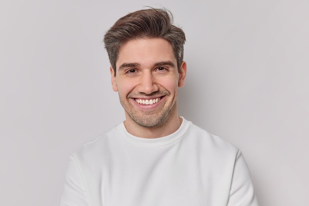 Free photo portrait handsome man with dark hairstyle bristle and toothy smile dressed in white sweatshirt feels very glad poses indoor pleased european guy being in good mood smiles positively emotions concept