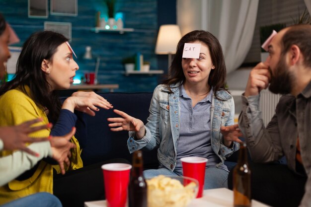 Portrait of happy woman with stickey notes on forehead playing guess who game enjoying spending time with friends socializing together during wekeend party at home. Friendship concept
