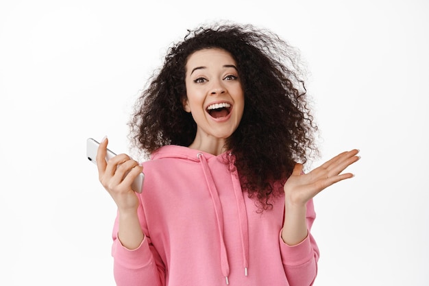 Free photo portrait of happy young woman use smartphone, rejoicing and shouting from joy, winning money on mobile phone, triumphing, winning on smartphone, standing against white background