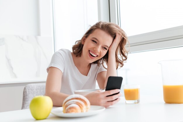 Портрет смех женщины в белой футболке, проверка новостей на мобильном телефоне, сидя за кухонным столом