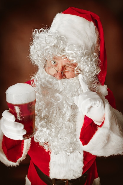 Foto gratuita ritratto di uomo in costume da babbo natale - con una lussuosa barba bianca, cappello di babbo natale e un costume rosso su sfondo rosso in studio con birra