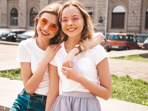 Бесплатное фото Портрет двух молодых красивых белокурых улыбающихся хипстерских девочек в модной летней белой футболке одевается.