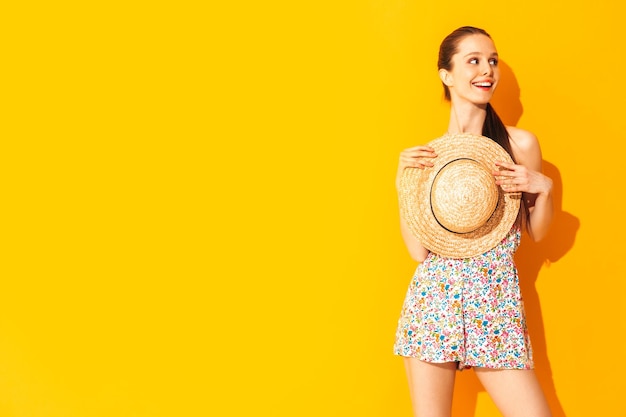 Бесплатное фото Портрет молодой красивой улыбающейся женщины в модном летнем платье беззаботная женщина позирует возле желтой стены в студии позитивная модель веселится в помещении веселая и счастливая держа шляпу в руках