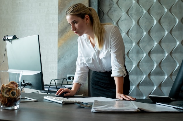 Бесплатное фото Портрет молодой женщины офисного работника, стоящей за офисным столом с документами, используя компьютер с уверенным и серьезным выражением лица, работающим в офисе