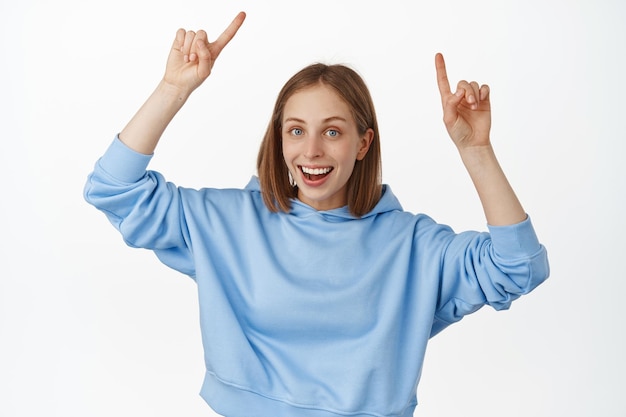 Бесплатное фото Портрет привлекательной молодой женщины, улыбающейся, указывающей пальцем на логотип, показывающей баннер со скидкой, рекламой вверх, стоящей в синей толстовке с капюшоном, рекомендую нажать на ссылку, белый фон.