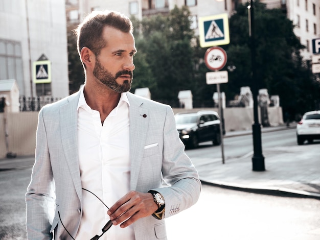 Бесплатное фото Портрет красивого уверенного в себе стильного хипстера ламберсексуальной моделисовременный мужчина в элегантном белом костюме модный мужчина позирует на фоне улицы в европейском городе на закате