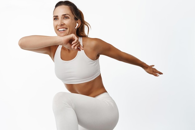 Бесплатное фото Портрет спортсменки, растягивающей тело, занимающейся фитнесом, поднимающей ногу и улыбающейся, бегущей, стоя на белом фоне