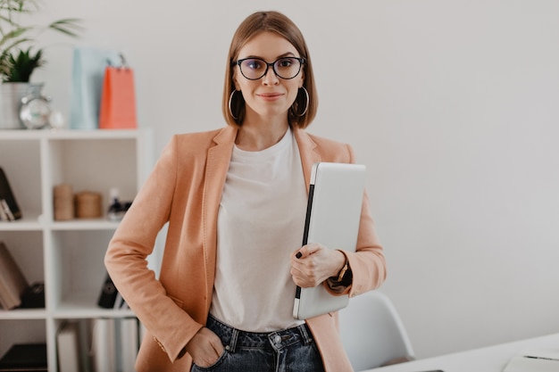 Бесплатное фото Портрет успешной деловой женщины в очках и в легкой куртке, улыбаясь против белого офиса.