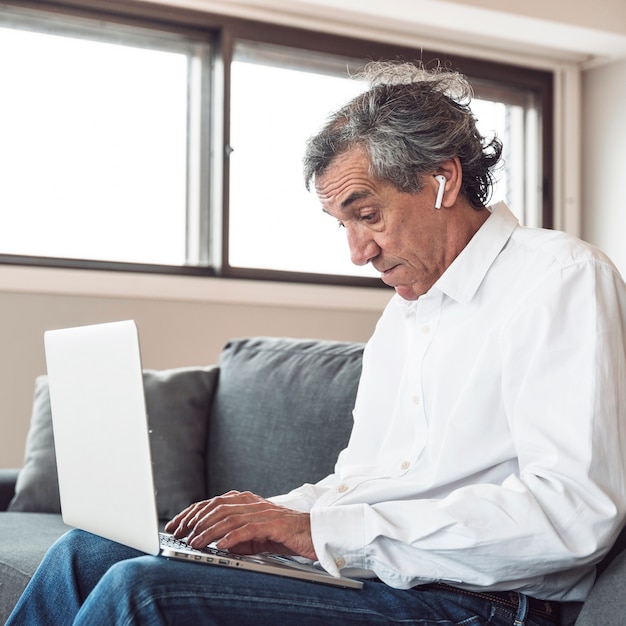 Free photo portrait of a senior man sitting on sofa wearing bluetooth earphone using laptop
