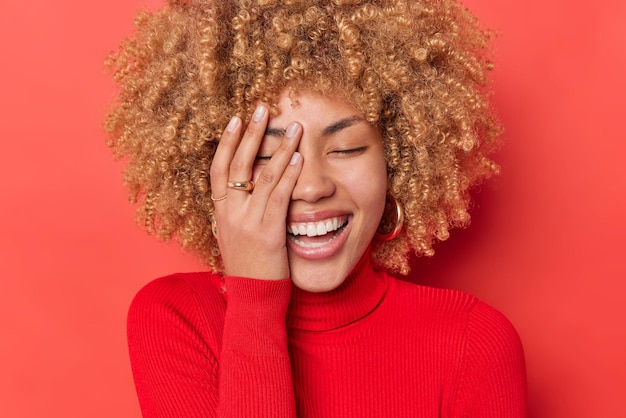 Free photo positive young woman with curly blonde hair giggles happily makes face palm expresses sincere emotions wears casual turtleneck isolated over red background. people happiness and joy concept.