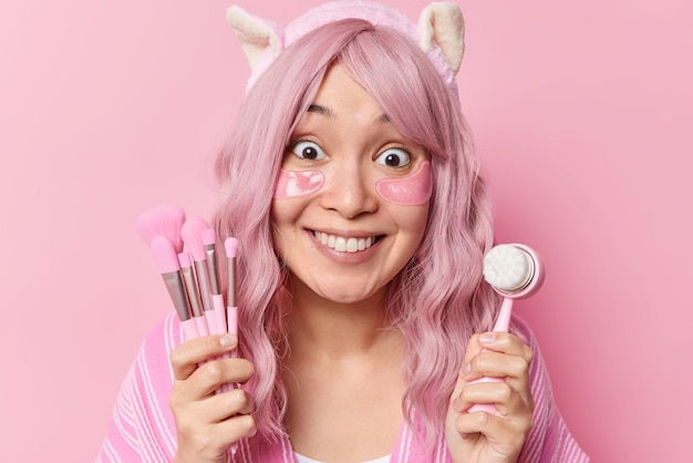 Free photo positive young asian woman with glad expression holds cosmetic brushes and face massager applies hydrogel patches has dyed hair isolated over pink background. skin care and wellness treatments