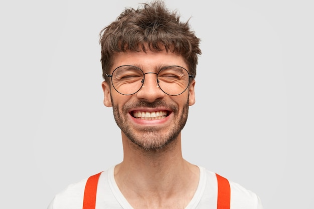 Free photo positive bearded man hipster smiles broadly, has pleased expression, laughs at something funny, closes eyes,