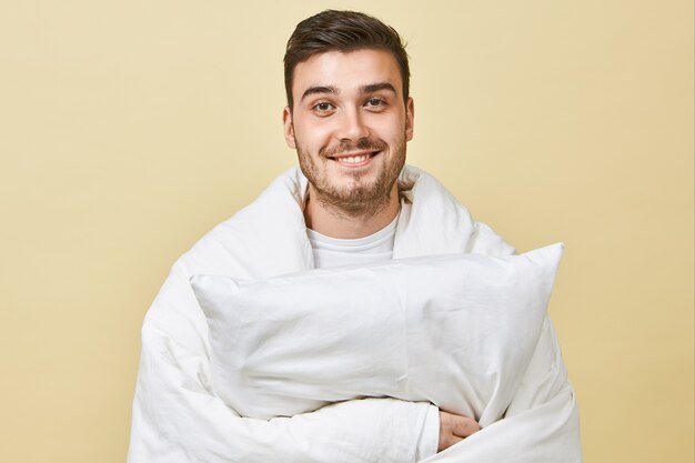 Positive cheerful young man with cute smile and unshaven face standing at blank wall, wrapped in white blanket, feeling overjoyed , recovering from cold, holding pillow, going to sleep in bed