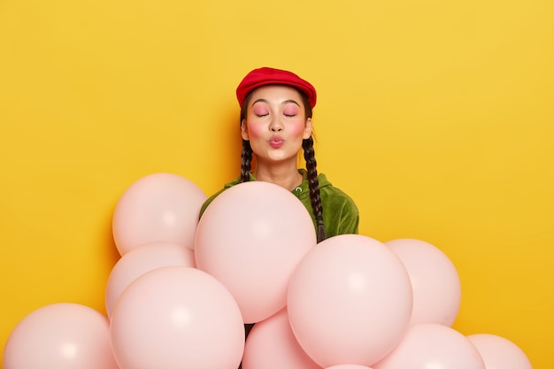 Бесплатное фото Довольная женщина с розовым макияжем, держит губы округлыми, хочет целовать гостей, благодарна за то, что пришли на вечеринку, стоит возле воздушных шаров, носит модный красный берет