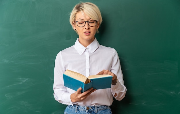 Бесплатное фото Довольная молодая блондинка учительница в очках в классе, стоя перед классной доской, указывая пальцем и читая книгу с копией пространства