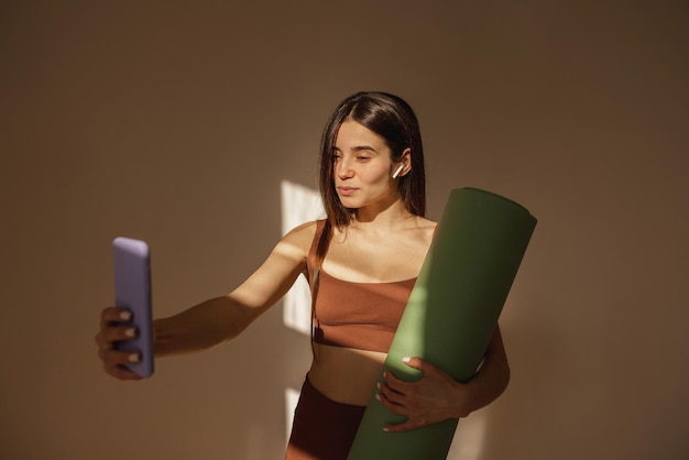 Бесплатное фото Довольно молодая кавказская дама с ковриком для фитнеса делает селфи на iphone в помещении. брюнетка в спортивной одежде наслаждается современными технологиями.