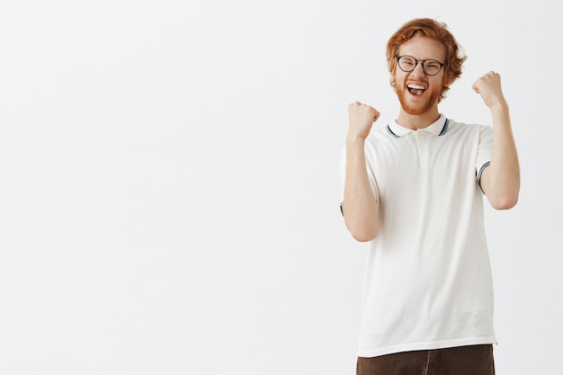 Бесплатное фото Радующийся и торжествующий бородатый рыжий парень позирует на фоне белой стены в очках