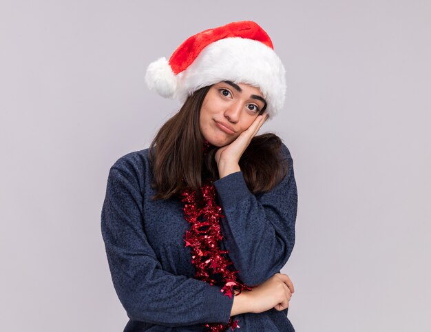 Sad young caucasian girl with santa hat and garland around neck puts hand on face and looks 