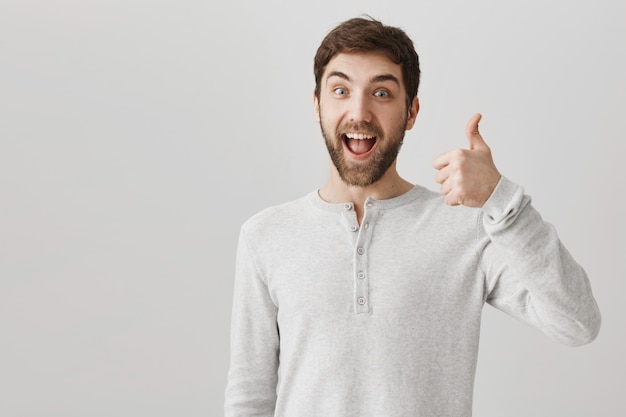 Бесплатное фото Довольный улыбающийся бородатый клиент-мужчина показывает палец вверх в знак одобрения