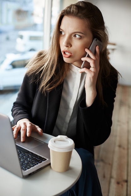 Бесплатное фото Европейская женщина, серьезно обеспокоенная, сидящая в кафе, пьющая кофе и работающая с ноутбуком, разговаривающая по смартфону, с тревогой глядя в сторону