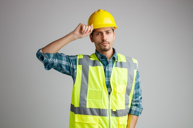Foto gratuita giovane ingegnere maschio serio che indossa casco di sicurezza e uniforme guardando la fotocamera mentre afferra il casco isolato su sfondo bianco
