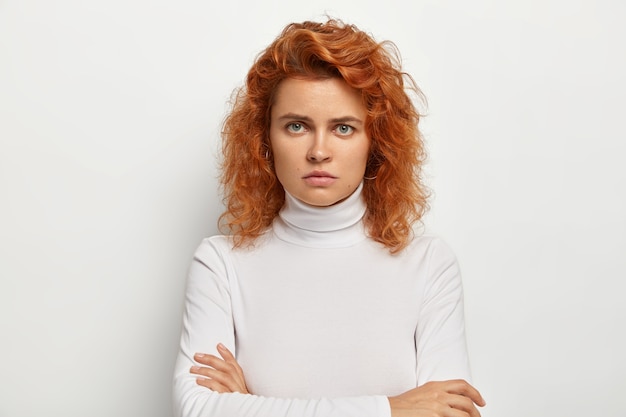 Free photo serious young lady with curly red hair, being dissatisfied with something, looks angrily, keeps hands crossed, wears white casual turtleneck, offended with dumb question, poses indoor alone.