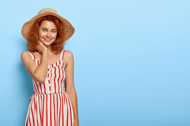 Бесплатное фото Выстрел счастливой рыжеволосой женщины, одетой в модный летний наряд, соломенную шляпу, с очаровательной улыбкой, естественной красотой, стоит у синей стены с пустым пространством для вашего продвижения.