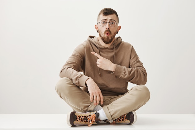 Free photo shocked man sit on floor, pointing finger left and looking concerned
