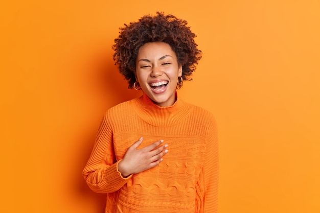 Free photo sincere emotions and positive feelings concept. happy pleased woman smiles broadly keeps hand on chest laughs at funny story dressed in casual sweater isolated over orange wall
