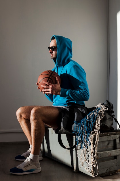Бесплатное фото Вид сбоку баскетболист, позирует в балахон и солнцезащитные очки с мячом близко к груди